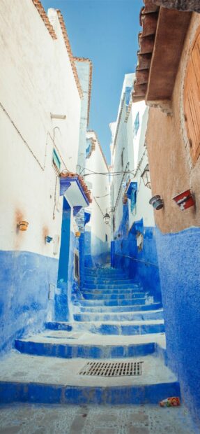 A narrow stairway painted in blue and white walls leads through a Moroccan town under a clear sky with hanging lanterns, 2K Mobile Wallpaper