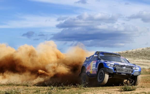A blue rally raid car races through a dusty field under a cloudy sky, 2K Desktop Wallpaper