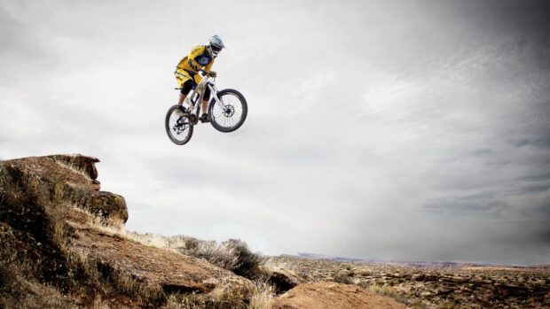 A Bmx rider in yellow gear is jumping over rocky terrain in a desert landscape, 4K Desktop Wallpaper