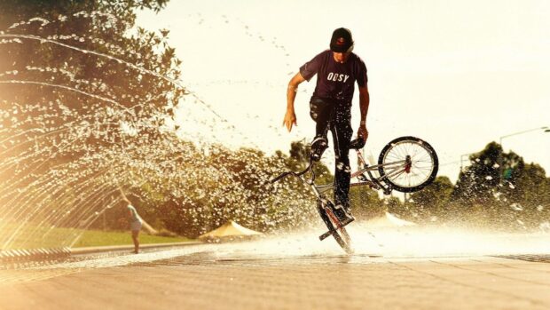 A BMX sports rider is doing a trick in front of water sprays at a park, HD Desktop Wallpaper