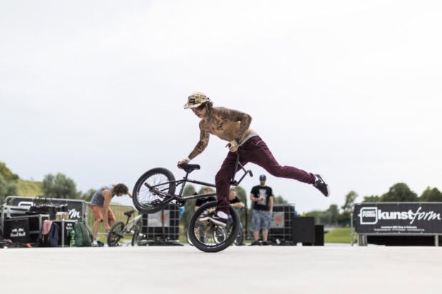 A tattooed BMX sports rider is balancing skillfully on one wheel outdoors, 2K Desktop Wallpaper