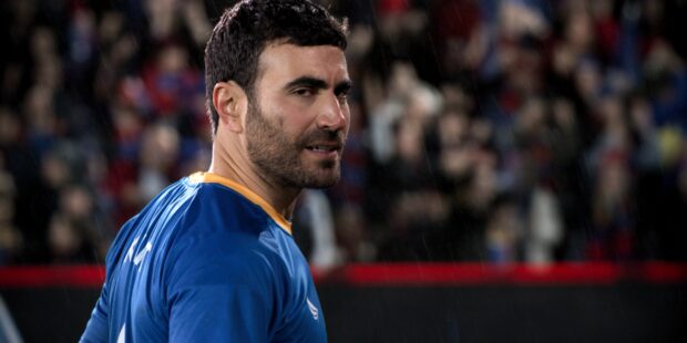 Brett Goldstein is wearing a blue sports shirt and looking over his shoulder in a rainy stadium, 2K Desktop Wallpaper