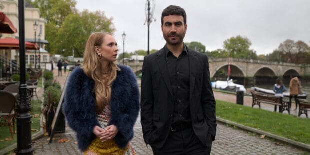 Brett Goldstein walking next to a woman in a blue fur coat near a river and bridge, 2K Desktop Wallpaper