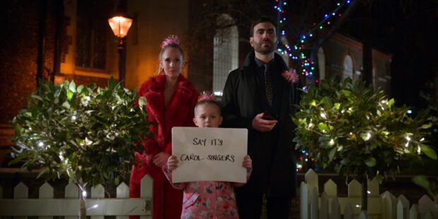 A man and woman stand behind a child holding a sign in a festive outdoor setting featuring Brett Goldstein, 2K Desktop Wallpaper
