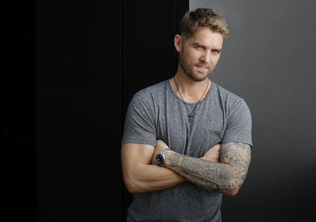 A man with tattooed arms and a watch stands with folded arms in front of a dark wall in Brett Young wallpapers, HD Desktop Wallpaper