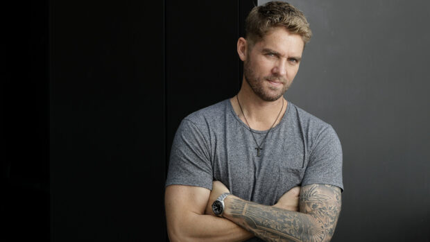 A man with tattooed arm and crossed arms wearing a gray shirt in a dark room, HD Desktop Wallpaper