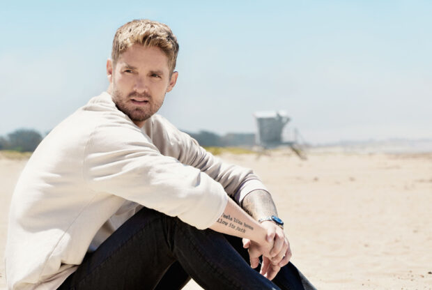 Brett Young sitting with a watch and tattoo on his arm at the beach, HD Desktop Wallpaper