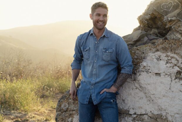Brett Young is standing outdoors near a rocky area with a smile on his face, 2K Desktop Wallpaper