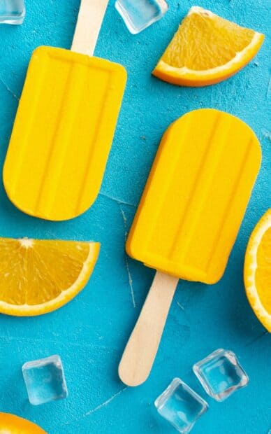 Bright yellow popsicle with orange slices and ice cubes on a blue surface shows refreshing summer treats, 2K Mobile Wallpaper