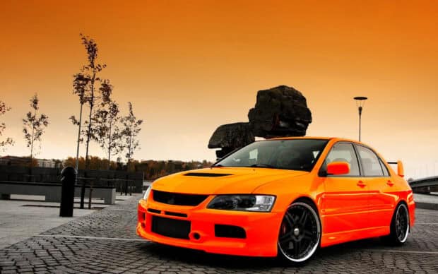 A bright orange Lancer Evo is parked on a cobblestone street under an orange sky near trees and a lamp post, HD Desktop Wallpaper
