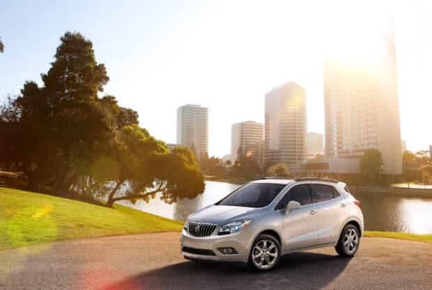A silver Buick Encore parked near a lake with modern buildings and trees in the background, HD Desktop Wallpaper