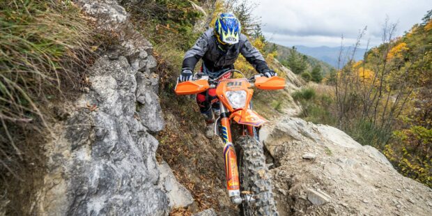 A rider on an enduro motorbike navigating a rocky mountain trail surrounded by autumn trees