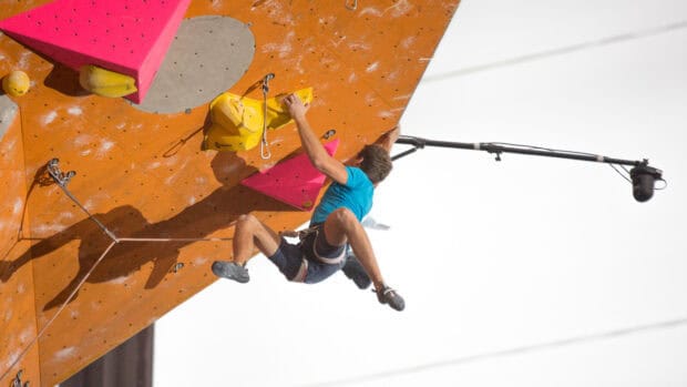A man in a blue shirt is climbing an orange indoor rock wall with colorful holds during a rock climbing competition, HD Desktop Wallpaper