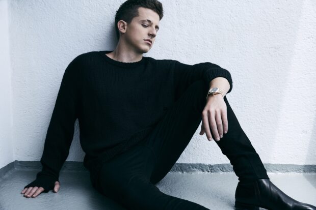 Charlie Puth is sitting on the floor leaning against a white textured wall wearing a black sweater and boots, HD Desktop Wallpaper