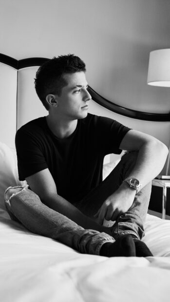 A young man in a black shirt sits thoughtfully on a bed in a black and white room, 4K Phone Wallpaper
