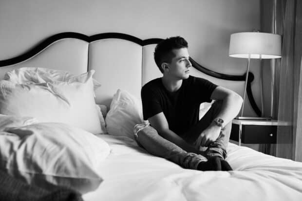 Charlie Puth sitting on a bed wearing a black shirt and jeans looking to the side, 2K Desktop Wallpaper