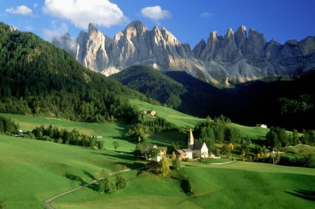 A peaceful village lies at the foot of towering rocky mountains on a sunny day with lush green fields, HD Desktop Wallpaper