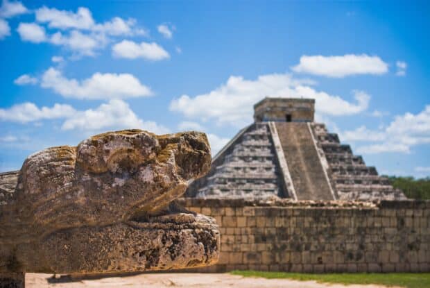 A stone sculpture stands with Chichen Itza pyramid in the background under a bright blue sky, 2K Desktop Wallpaper