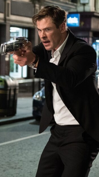 Chris Hemsworth is aiming a gun with intense focus in a dark suit on a city street, 4K Phone Wallpaper