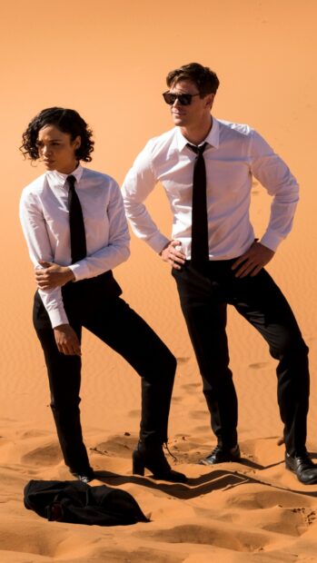 Chris Hemsworth stands beside a woman in Men In Black outfits on the desert sand, HD Mobile Wallpaper