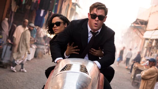 Chris Hemsworth rides a futuristic vehicle with a partner wearing sunglasses in a busy market, 4K Desktop Wallpaper