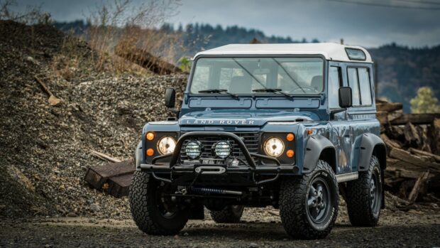 A classic Land Rover Defender is shown parked on rocky terrain with headlights on, HD Desktop Wallpaper