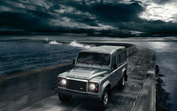 A Land Rover Defender drives along a winding coastal road with stormy clouds above, 2K Desktop Wallpaper