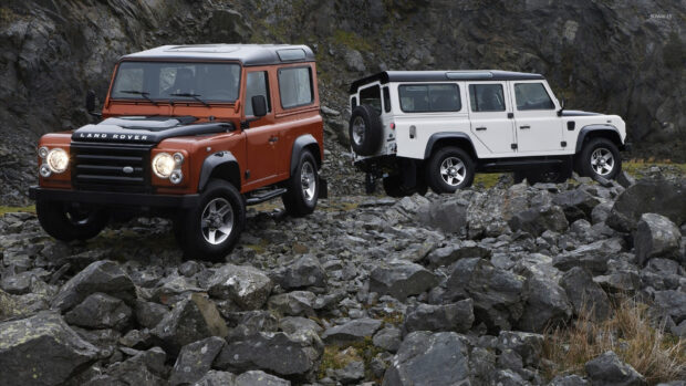 Two Land Rover Defender vehicles are parked on rough rocky ground near a mountainous area, HD Desktop Wallpaper