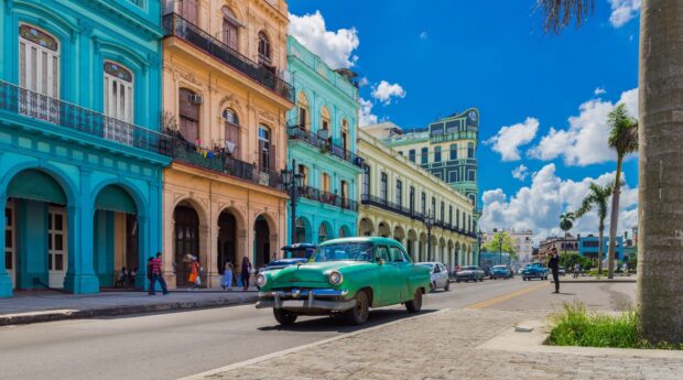 A green vintage car drives past colorful buildings under a blue sky in Havana Cuba, HD Desktop Wallpaper