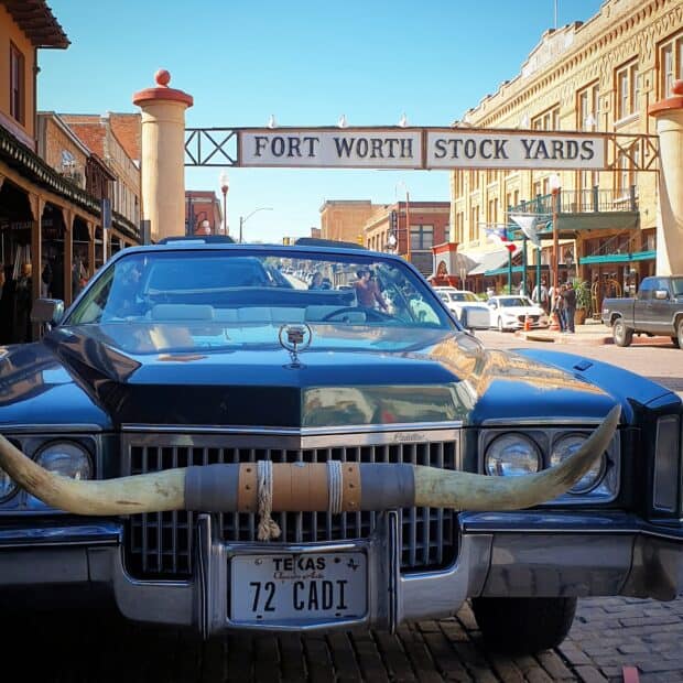 A classic car with longhorn hood ornament is parked under the Fort Worth Stock Yards sign, HD Mobile Wallpaper