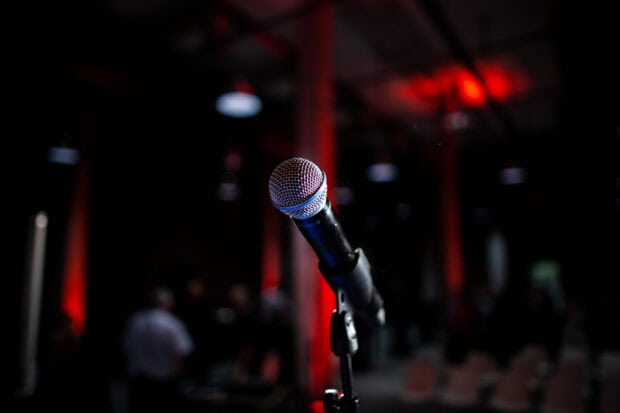 A classic microphone stands ready in a dimly lit room with red accents and blurred people, 2K Desktop Wallpaper