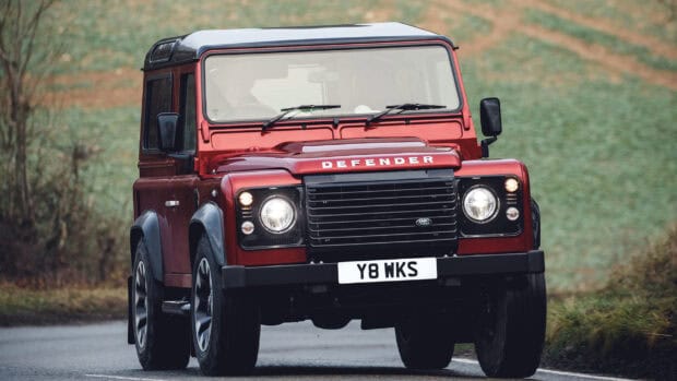 A red Land Rover Defender is driving on a country road amid green fields, 2K Desktop Wallpaper