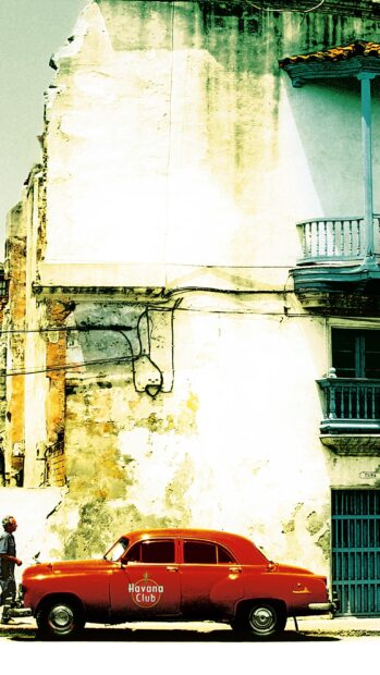 A classic red car is parked by an old wall in Havana Cuba with a person walking nearby, 2K Mobile Wallpaper