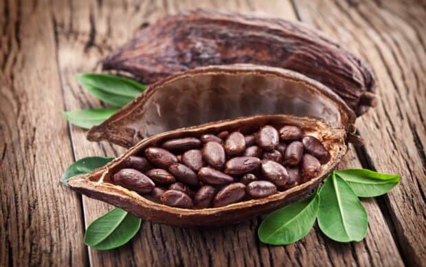Cocoa beans inside a cracked cocoa pod with green leaves on wooden surface, 2K Desktop Wallpaper