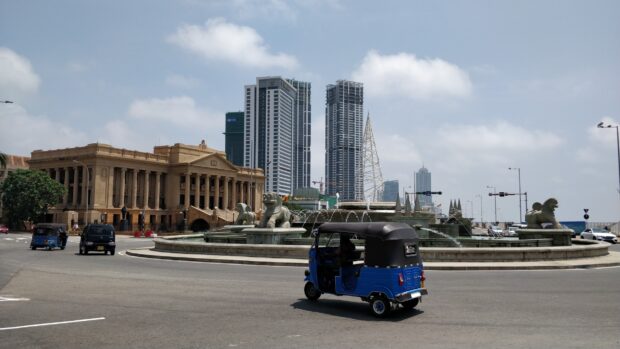 A blue tuk tuk drives near a decorative fountain and modern buildings in Colombo Sri Lanka, HD Desktop Wallpaper