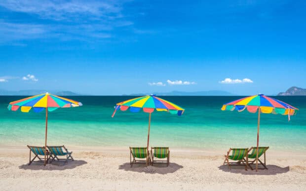 Three striped umbrellas with chairs are set on the sandy beach near the blue ocean, 2K Desktop Wallpaper