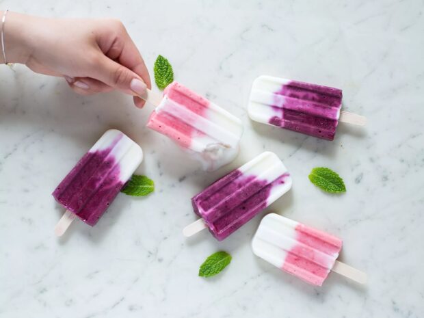 A hand holding a pink and white popsicle among five vibrant purple and white popsicles with fresh mint leaves on marble, HD Desktop Wallpaper