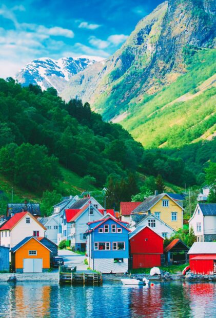 A vibrant village sits by the calm water surrounded by green hills and snowy mountains in Norway wallpapers, HD Mobile Wallpaper
