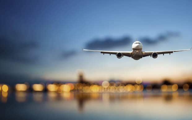 A commercial aircraft is flying low over a cityscape with bright lights reflecting on the water during dusk, 2K Desktop Wallpaper