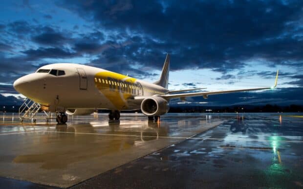 A commercial aircraft with yellow accents is parked on the wet runway under a cloudy evening sky, 2K Desktop Wallpaper