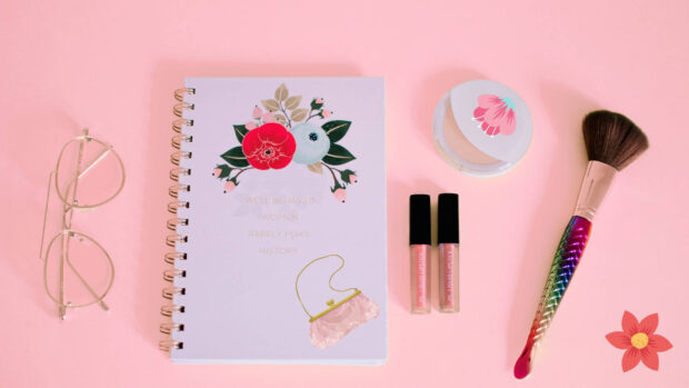 A makeup brush and two lipsticks sit next to a floral notebook and glasses on pink, HD Desktop Wallpaper
