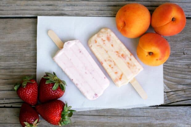 Two creamy popsicles surrounded by fresh strawberries and apricots on a wooden surface, HD Desktop Wallpaper