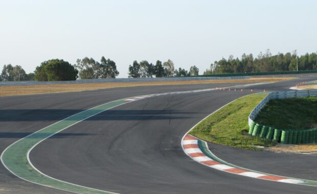 A curved race track shows clear asphalt and safety barriers under a blue sky with trees, 2K Desktop Wallpaper