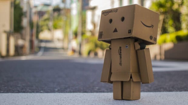A cardboard robot doll is standing on a street with a blurred urban background featuring Amazon logos, HD Desktop Wallpaper