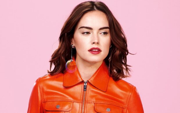 Daisy Ridley wears an orange leather jacket and colorful earrings in front of a pink background, HD Desktop Wallpaper