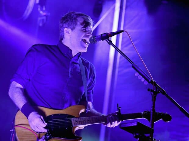 A musician from Death Cab For Cutie sings and plays guitar on stage with purple lighting, 2K Desktop Wallpaper