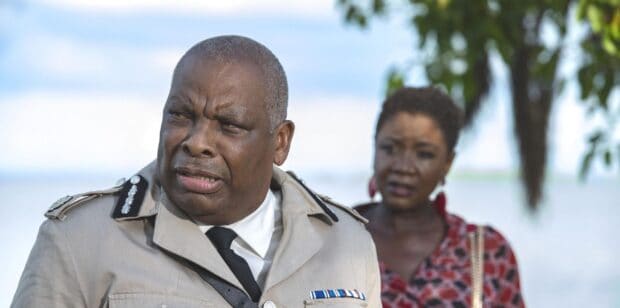 A scene from Death In Paradise TV series showing two characters outdoors near water, 2K Desktop Wallpaper