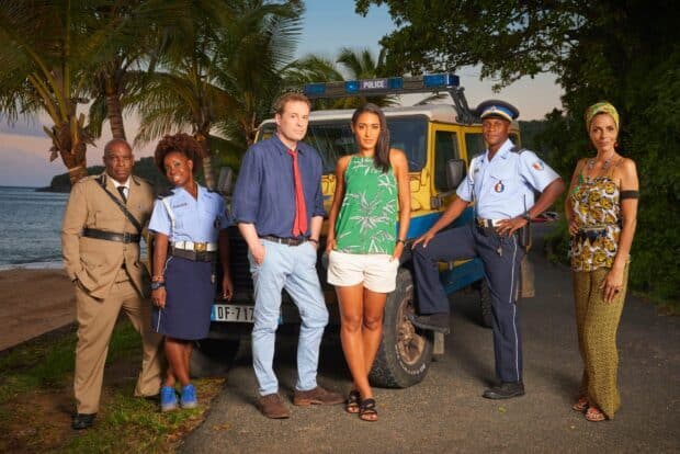 The cast of Death In Paradise tv series stands by a police vehicle on a tropical coastal road, HD Desktop Wallpaper
