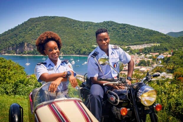 Two police officers from Death In Paradise tv series pose by a motorcycle with a tropical landscape, HD Desktop Wallpaper