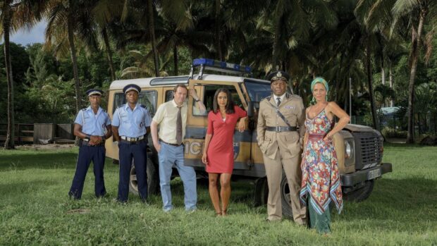 The Death In Paradise tv series cast stands outdoors near a police jeep on a grassy field, HD Desktop Wallpaper